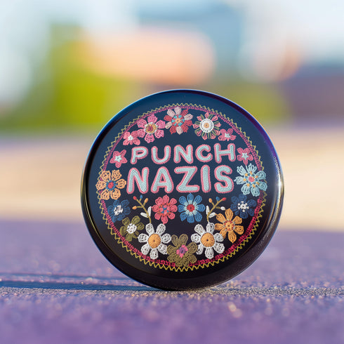Round pinback metal button featuring an embroidered-style design on a black background. The center showcases bold, stitched-textured lettering in red, blue, and white that reads "PUNCH NAZIS." Surrounding the text is an intricate, folk-inspired floral pattern with multicolored flowers in shades of pink, orange, blue, and white, arranged symmetrically. A decorative zigzag border frames the edge of the button. The backside is silver metal with a secure pin closure.